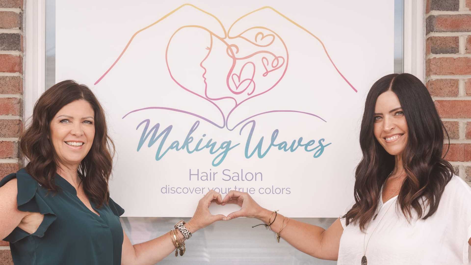 Two women forming a heart shape with their hands in front of "Making Waves Hair Salon" sign.