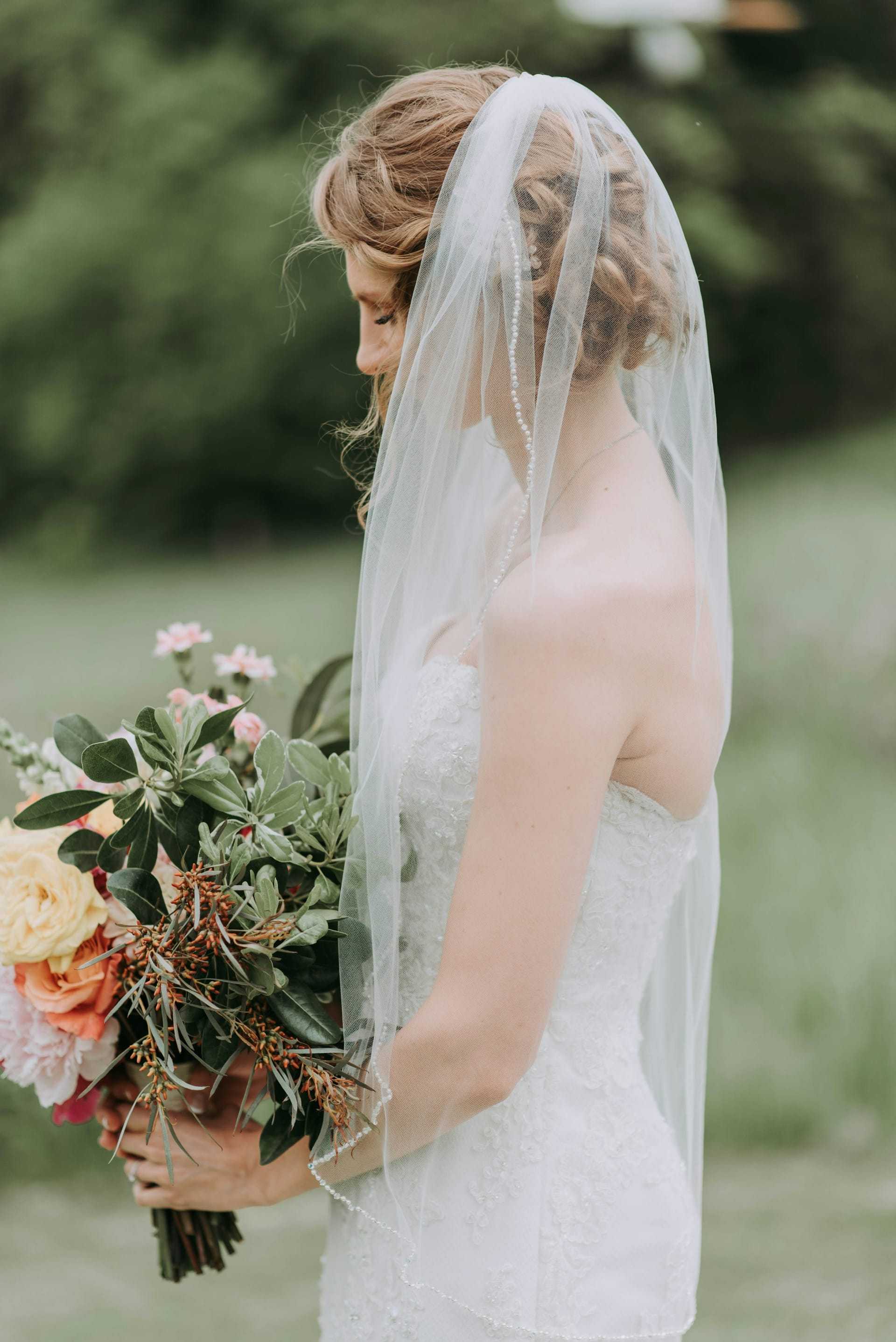 Bride in white gown holding a bouquet, looking down outdoors with a veil over her face.