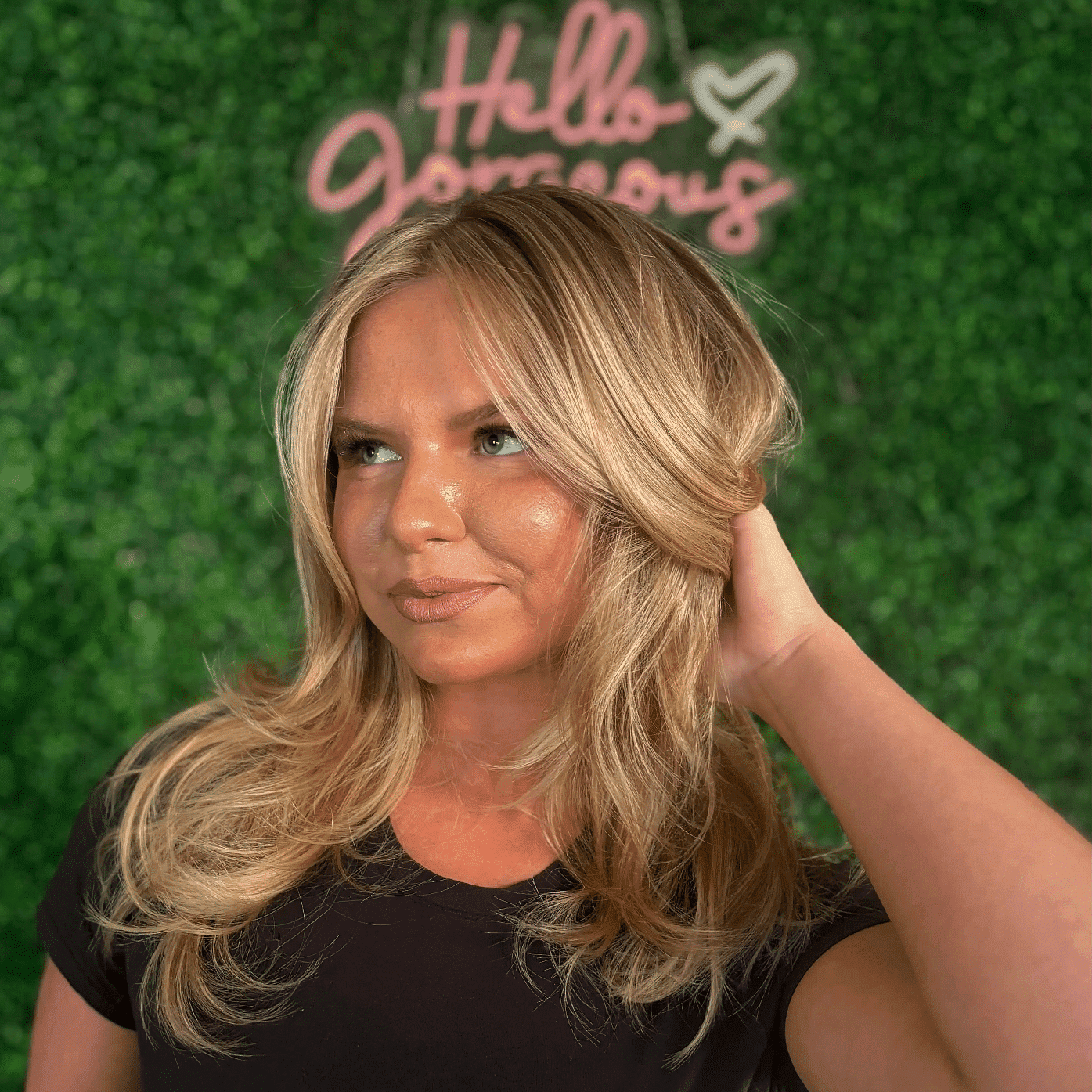 Woman with blonde hair in black top poses in front of greenery wall with "Hello Gorgeous" sign.